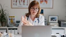 A doctor with glasses sits at a desk and speaks during a telehealth visit.