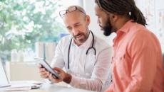 A male doctor sits with another man and they are looking at SDOH data on a mobile device.